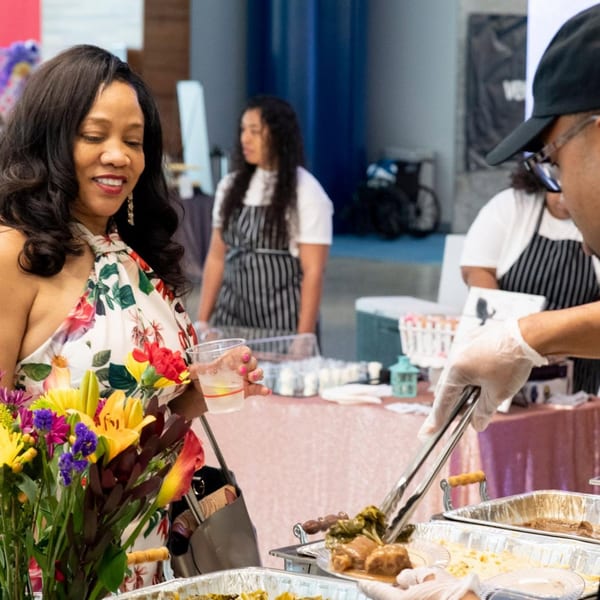 Woman in floral dress smiling while a server plates food at a buffet event.