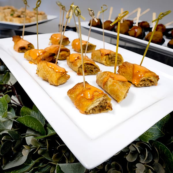 Crispy spring rolls with savory filling, drizzled with orange sauce and skewered on a white platter.