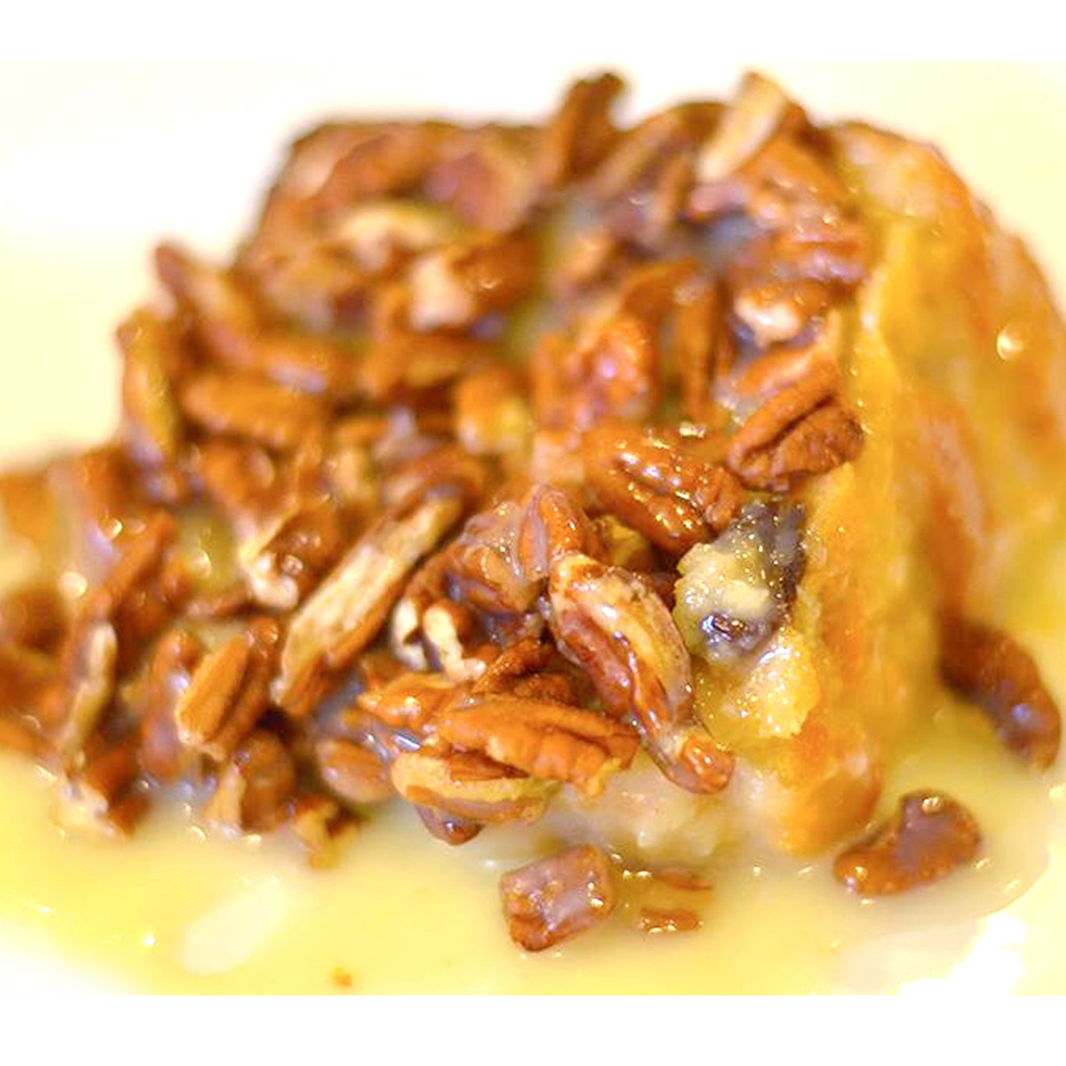 Golden dessert topped with caramelized pecans and mixed nuts, drizzled with syrup on a white plate.