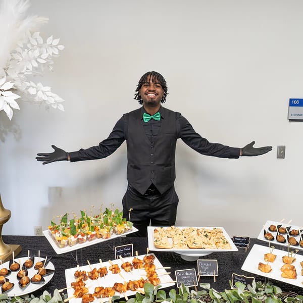 Smiling server in a green bow tie presents a buffet table laden with various appetizers.