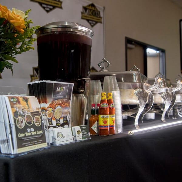 Buffet table with chafing dishes, a drink dispenser, hot sauces, and MKTS catering brochures.
