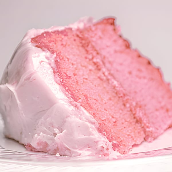 A close-up of a slice of pink strawberry cake with light pink frosting on a white plate.