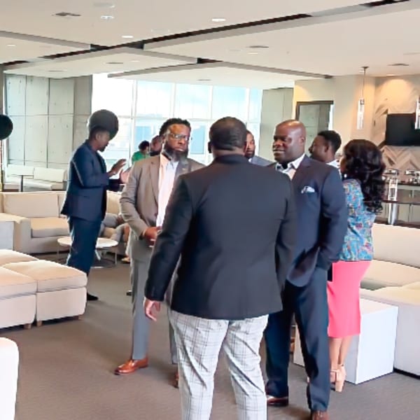 Formally dressed people conversing in a bright, modern lounge with white sofas and large windows.