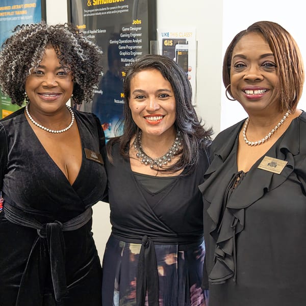 Three smiling women in black dresses and necklaces pose in front of career information posters.
